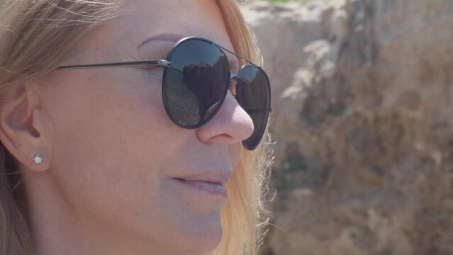 Close-up face of confident carefree blond woman in sunglasses looking away in sunshine. Closeup portrait of relaxed satisfied female Caucasian tourist in sunlight on summer Cyprus