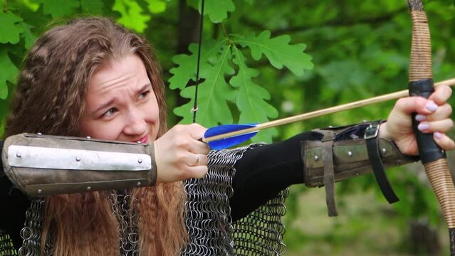 Smooth camera movement, selective focus, portrait of a young woman in chain mail and bracers drawing a bowstring and aiming a bow, against the backdrop of a summer forest.