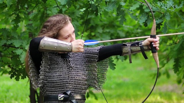 Smooth camera movement, selective focus, portrait of a young woman in chain mail and bracers drawing a bowstring and aiming a bow, against the backdrop of a summer forest.