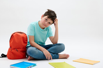 Puzzled pensive doubtful teenager, school child boy, scratching his head, looking at the camera, sitting next to arranged textbooks and orange backpack on isolated white background. Education concept - obrazy, fototapety, plakaty