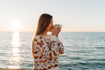 Fototapeta na wymiar Young beautiful girl in cozy sweater drinking coffee while enjoying winter sun on seaside shore during mild sunset. Cute attractive woman enjoying cup of tea with autumn ocean sun trail background.