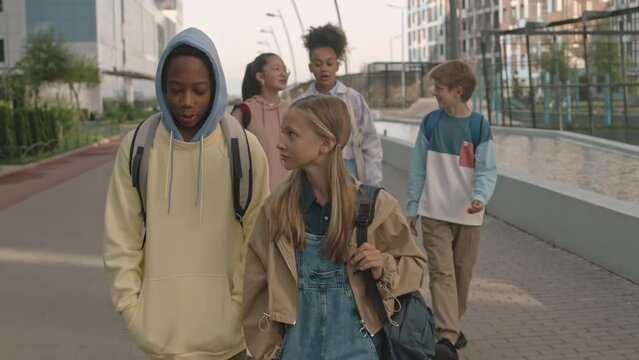 Tracking shot of multiracial group of preteen schoolkids chatting while walking together on sidewalk in summerTracking shot of multiracial group of preteen schoolkids chatting while walking together o