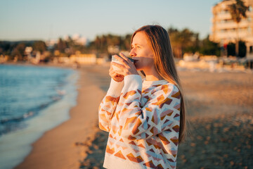 Young beautiful girl in cozy sweater drinking coffee on a winter seaside shore while taking...
