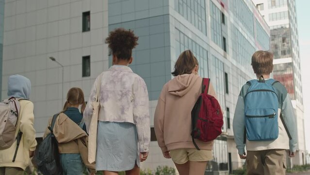 Tracking medium back view shot of group of diverse preteens with school backpacks walking outdoors in city on sunny day