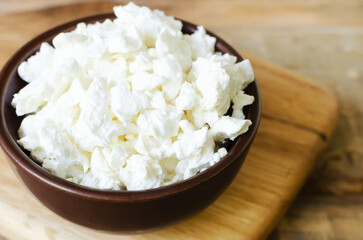 Organic cottage cheese in a brown ceramic bowl on a wooden cutting board on a wooden table. The concept of home-made food. Selective focus. Horizontal orientation.