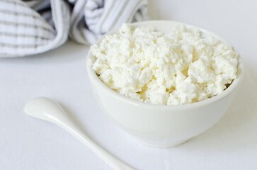 Organic cottage cheese in a white ceramic bowl with a teaspoon on a white tablecloth. Healthy breakfast. The concept of homemade food. Selective focus. Horizontal orientation.