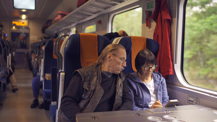 Elderly couple travel by train, the lady is holding a mobile phone in her hand, both are looking at the smartphone and talking with each other