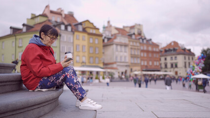 Elderly lady sits on the steps drinking coffee and using a smartphone in the historic center of an old European city. Palace Square, Warsaw Old Town