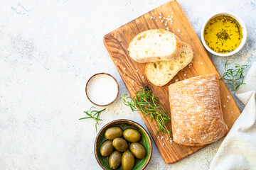 Ciabatta bread on wooden board with olive oil, olives and herbs. Top view on white table. - obrazy, fototapety, plakaty