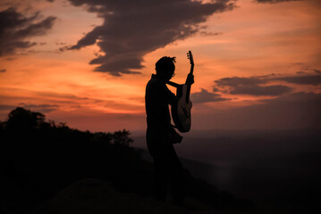 A silhouette man is playing the guitar in the mountains. as the sun was rising in the morning. The light of the sky has a beautiful orange color.