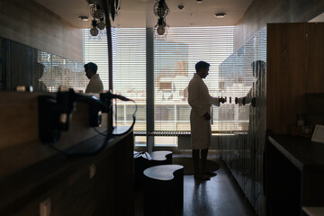 Silhouette of unrecognizable man in white robe after spa treatment at wellness center opening locker to change clothes in dark dressing room with mirrored walls, standing by window.