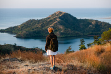 A young girl, blonde, with a backpack, stands on top of a mountain, rear view, overlooking a beautiful panorama of the islands and bays in Indonesia.