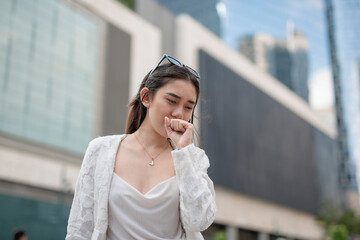 A young asian woman coughs while walking around the city. Urban air pollution issues or allergens.