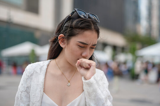 A young asian woman coughs while walking around the city. Urban air pollution issues or allergens.