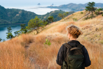 A young girl, blonde, with a backpack, stands on top of a mountain, rear view, overlooking a beautiful panorama of the islands and bays in Indonesia.