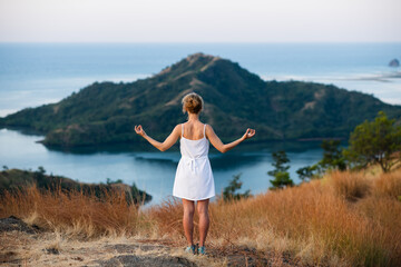 A beautiful, young girl of European appearance, with blond, curly hair, in a white dress, meditates, against the backdrop of a beautiful landscape of islands and bays.