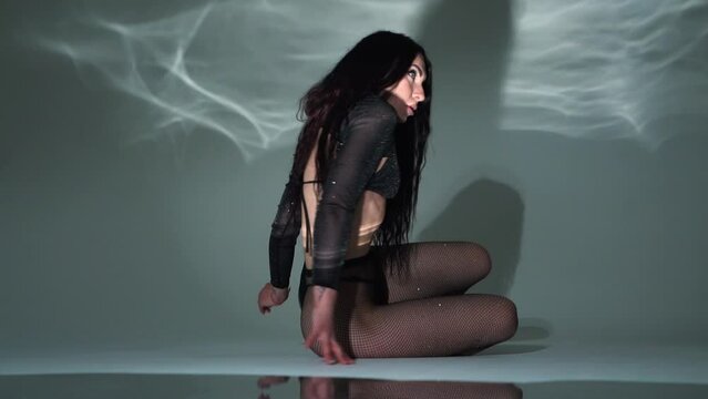 A brunette girl in black underwear with a beautiful athletic figure poses in a dark studio with light, lies on the floor in pantyhose