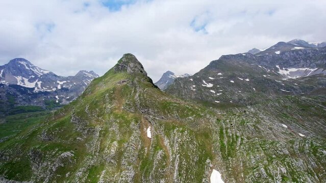 A mountain peak around which the road twists, shot from a drone in spring, behind which the highest mountain peaks with remnants of snow can be seen.