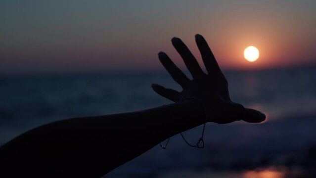 Silhouette of female Caucasian hand closing red sun in darkness of sunset over Mediterranean sea. Closeup unrecognizable woman opening star gesturing stretching arm to sundown