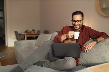 Smiling man holding a cup of coffee, using a laptop, watching something online.