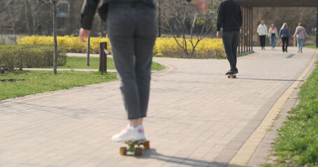 Young male and girl on skateboards ride along the path in city park with green grass. Unrecognizable, rear view, spring daytime. People rest, walk in the park.