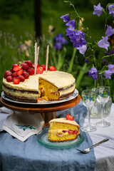 Homemade strawberry Birthday cake with three candles and cut out slice on wooden cake stand on round table in blooming garden.