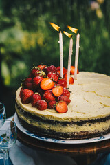 Homemade strawberry Birthday cake  with three lit candles on wooden cake stand on round table in blooming garden.