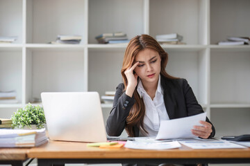 A stressed and serious young Asian businesswoman focuses on working on her business financial reports at her desk in the office.