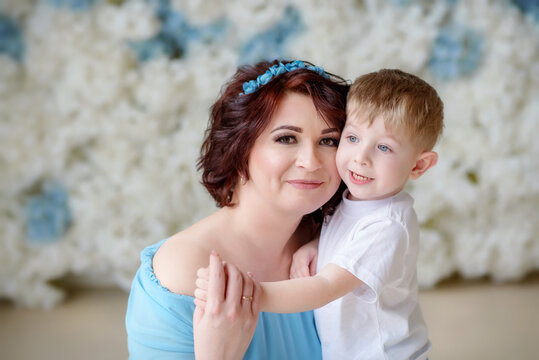 Mom hugs her son. Happy family in a bright room. Tenderness. A woman in a beautiful long blue dress. Family history. Younger son. A woman and her son at a photo shoot. Portrait of mother and son