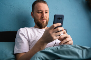 Portrait of smiling young freelance businessman using mobile phone checking social media network sitting on bed at home. Online digital communication