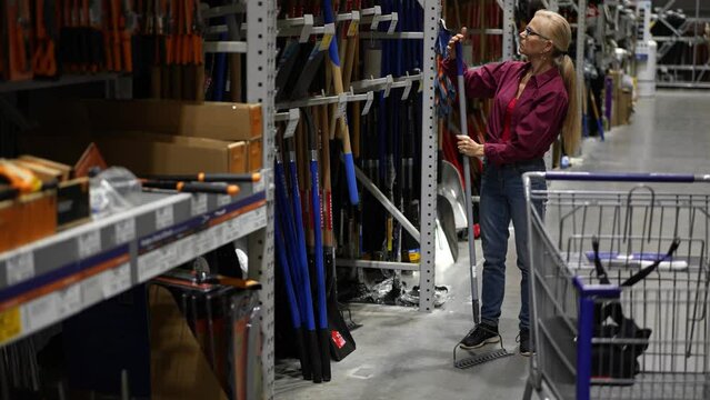 Attractive happy mature woman looking at rake garden tool in a large hardware store.