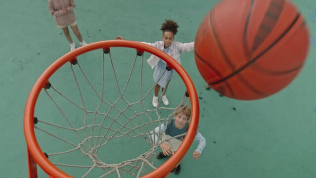 High angle slowmo with selective focus on basket of four multiethnic schoolkids standing at outdoor court and trying to throw basketball in basket one by one