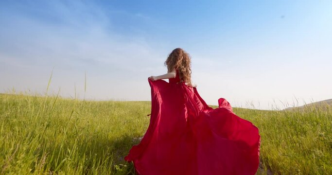 Caucasian red-haired woman in long red dress gently confidently rushes across field in dawn rays. Woman enjoys beautiful spectacle that opens her soul. Woman feels eternal love for freedom and beauty.