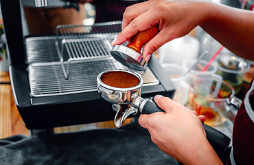 Hand of a barista holding a portafilter and a coffee tamper making an espresso coffee. Barista presses ground coffee using a tamper in a coffee shop