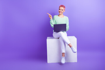 Full size photo of charming cheerful girl sit podium cube use netbook indicate finger empty space offer isolated on violet color background