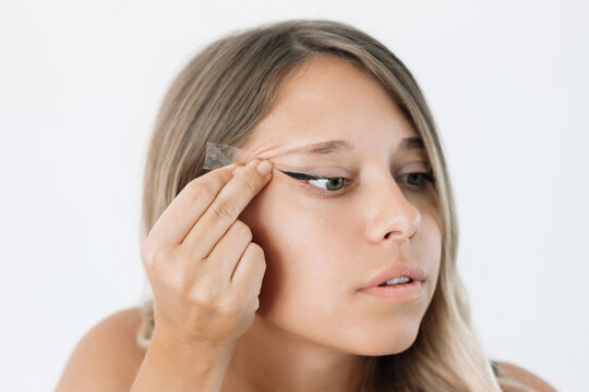 Young caucasian blonde woman wants to change her eye shape pulling eyelids to the side with hand using transparent tape isolated on white background. Plastic surgery, blepharoplasty concept. Close up