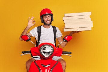 Young happy man in red helmet posing on red scooter against yellow backdrop, holds pack of pizza boxes in one hand, shows ok with other hand, delivery service concept, copy space