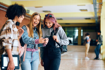 Cheerful high school friends using smart phone during break in hallway.