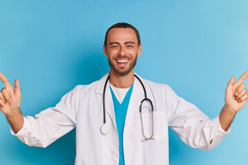 Happy male doctor in medical white coat posing against blue background with wide smile on his face, holding index fingers up, professional people concept, copy space
