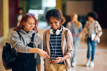 Happy black high school student and her friend using cell phone while walking through hallway at...
