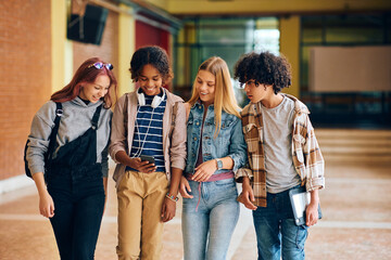 Happy teenage friends using smart phone while walking through high school hallway.