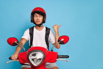 Oriental courier sitting on red scooter holding handle bar with one hand and pointing aside with...