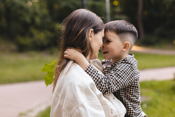 Happy mom holding son boy in arms, hugging kid with noses touch, smiling, enjoying warm moment. Younger mum carrying kid and dancing in the park. Motherhood, adoption, family concept. Focus on mom ear