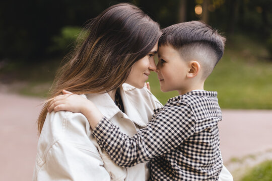 Happy mom holding son boy in arms, hugging kid with noses touch, smiling, enjoying warm moment. Younger mum carrying kid and dancing in the park. Motherhood, adoption, family concept.