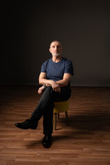 an ordinary adult man with a beard is sitting on a chair in the studio, dark background, selective...