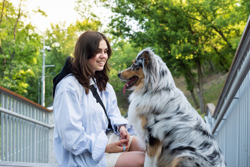 Portrait of a happy young woman and her aussie dog in a park. Blue merle australian shepherd dog in urban park area next to female owner, walking pets in the city