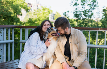 Young happy woman and man hug their cute aussie dog in a park. Blue merle australian shepherd dog in urban park area next to owners, taking pets for a walk in the city