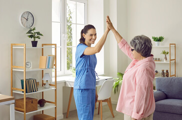 Patient with doctor. Funny and satisfied senior woman gives high five to young nurse who visited her at home. Young smiling woman in nurse's uniform supports elderly patient during medical home visit.