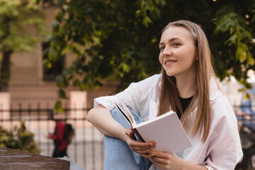 Pleased blonde woman sitting on bench and reading book in park. Dreaming girl studying or read book...