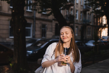 Outdoor photo of smiling happy woman with wonderful smile holding summer cocktail on blur city background in sunny warm evening. Girl walk in town and enjoy of refreshment lemonade. Closed eyes.